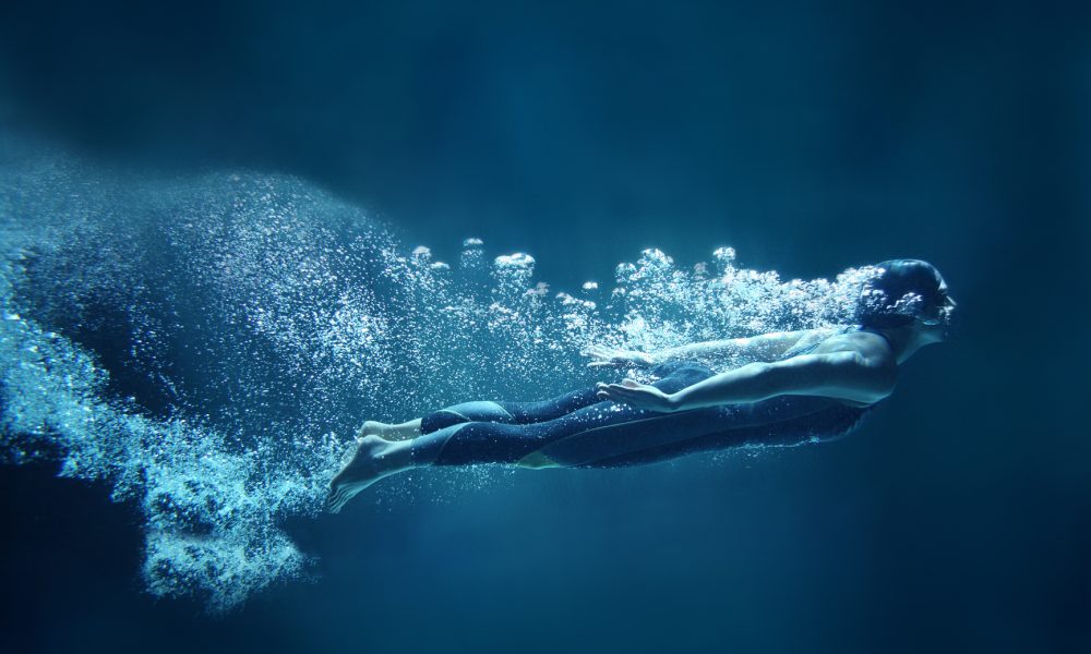 Athlete is dressed in a professional black swimwear. She is swimming horizontally like she was flying. She has two hands together along the body. She is looking ahead.  Behind her body you can see a lot of air bubbles. The background is dark blue- like open water. This is a square format photography.